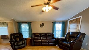 A living room with a sectional sofa, two leather armchairs, a ceiling fan, and a window with curtains. - A little bit of country (Aztec)