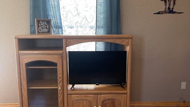 A wooden entertainment center with a TV, a decorative sign, and a window with curtains. - A little bit of country (Aztec)