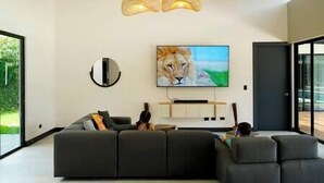 A modern living room with a large black sectional sofa, a flat-screen TV mounted on the wall, and a large window providing a view of the outdoors. - Casa Arena Volcano View Villa (Fortuna)