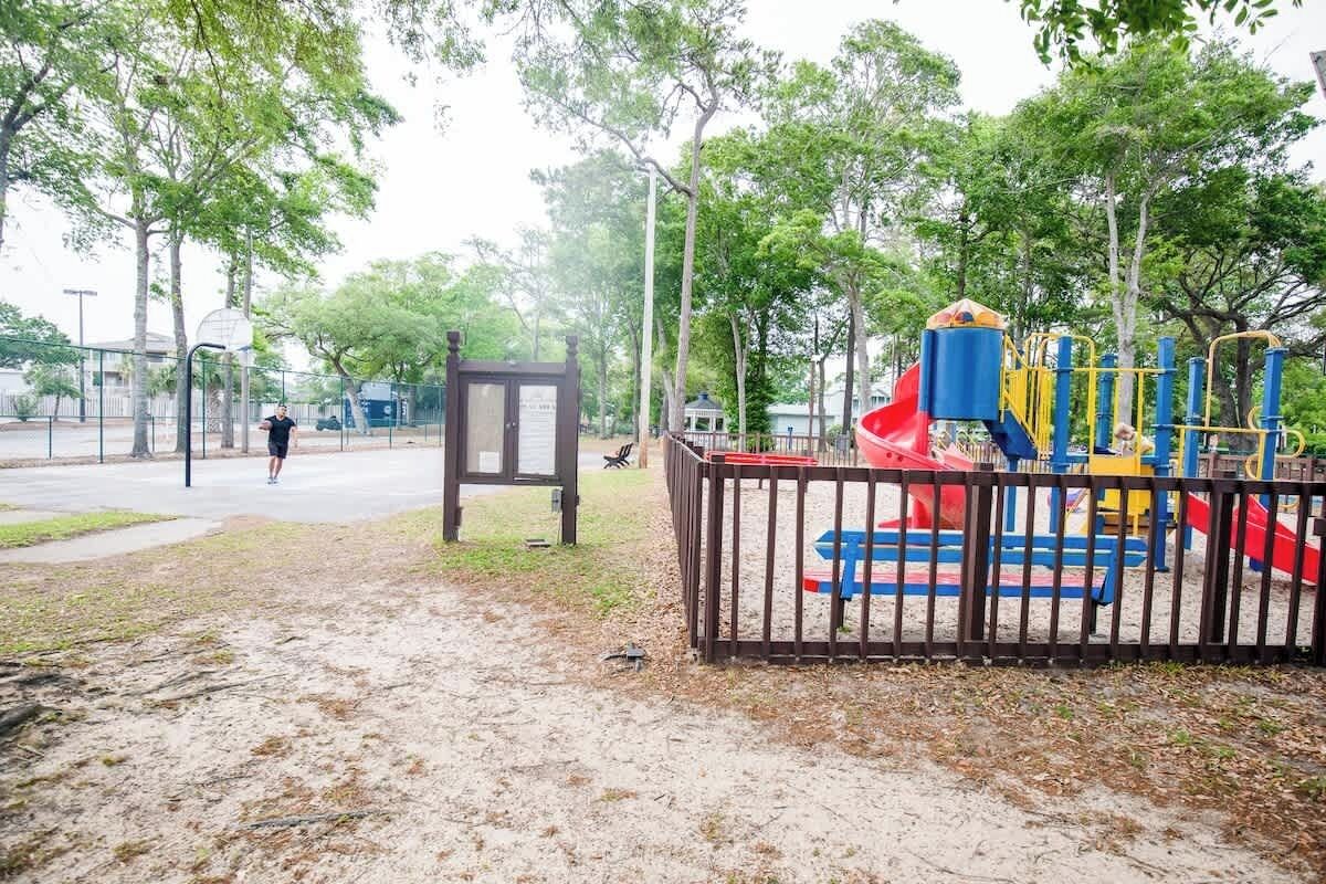 A playground with a slide and climbing structures, surrounded by trees and a basketball court.