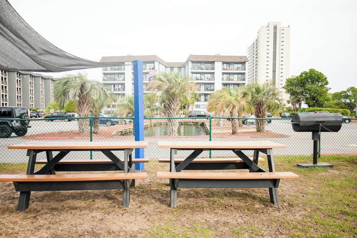 Deux tables de pique-nique, un poteau bleu, une clôture et un parking avec des voitures, ainsi qu’un bâtiment en arrière-plan.