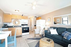 A modern kitchen with wooden cabinets, a dining area with a white table and blue chairs, a living room with a dark green sofa, and a ceiling fan.