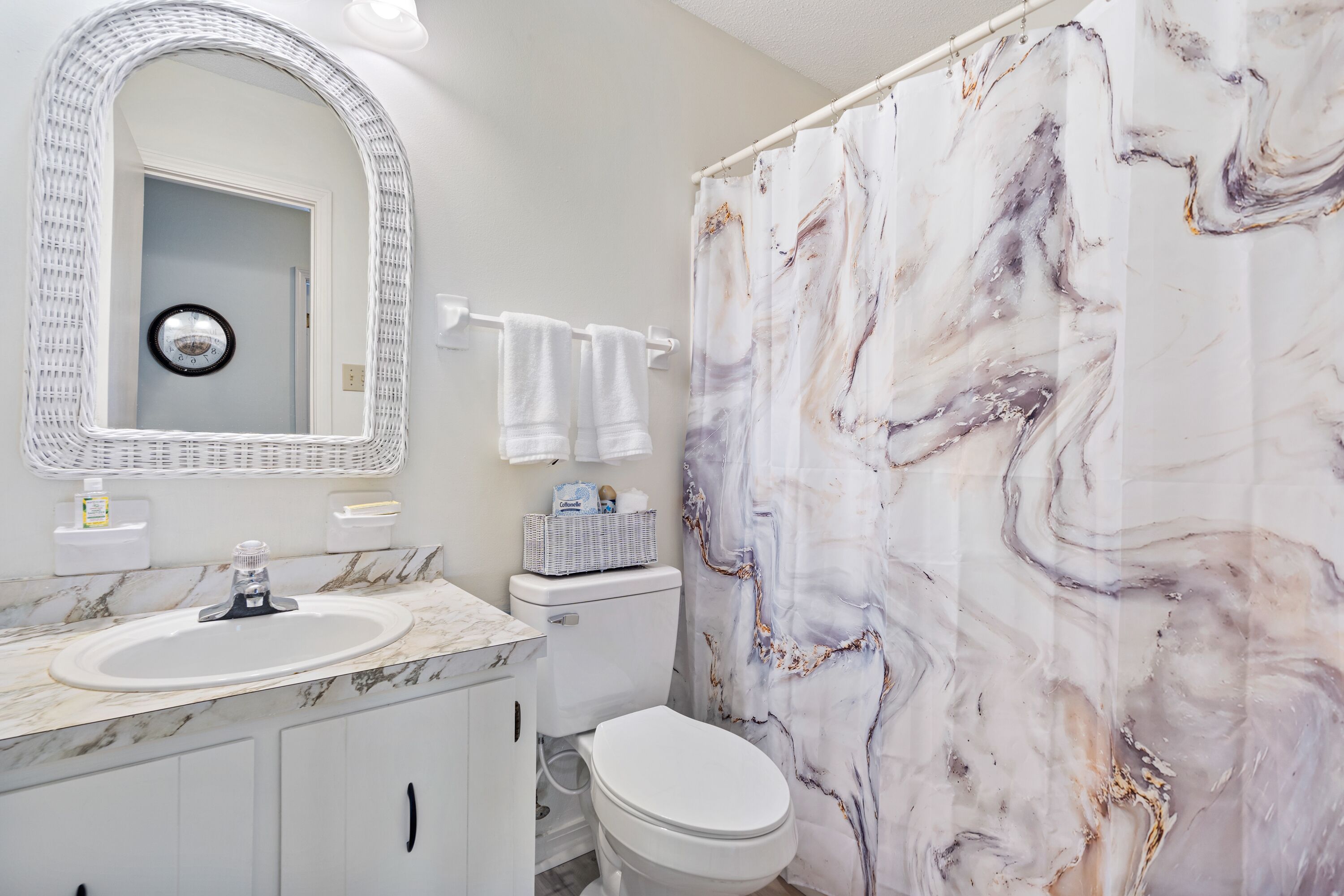 A bathroom with a marble sink, a toilet, a shower curtain with a marble pattern, a white wicker chair, and a wall-mounted mirror.