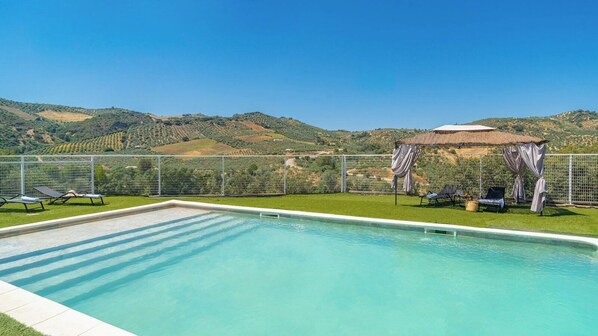 A swimming pool with a view of green hills and a fenced area with lounge chairs.