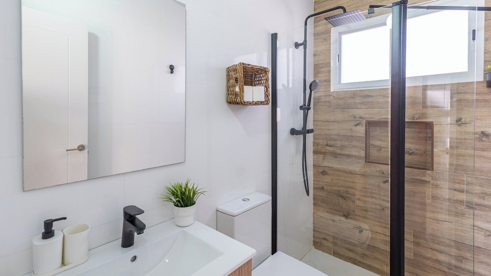 A modern bathroom with a large mirror, a white sink, a black faucet, a small potted plant, a shower with a glass door, and a wicker basket on the wall.