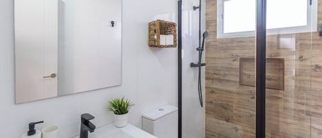 A modern bathroom with a large mirror, a white sink, a black faucet, a small potted plant, a shower with a glass door, and a wicker basket on the wall.