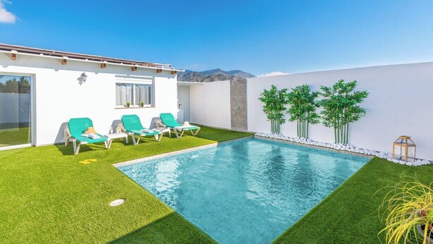 Une piscine bordée de gazon synthétique vert, un bâtiment blanc et, au loin, une vue sur les montagnes.