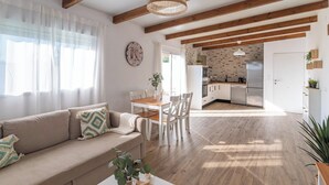Un salon avec un canapé, une table à manger entourée de chaises, une cuisine à l’arrière-plan, des poutres en bois au plafond et une horloge au mur.