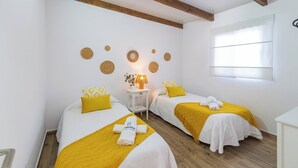 Une chambre d’hôtel avec deux lits, chacun recouvert d’une couvre-lit jaune et agrémenté d’oreillers, une petite table de chevet avec une lampe, et une fenêtre avec des stores.
