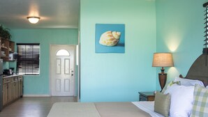 Quarto com cama, mesa de cabeceira, luminária, parede com quadro, porta e área de cozinha com armários e janela.