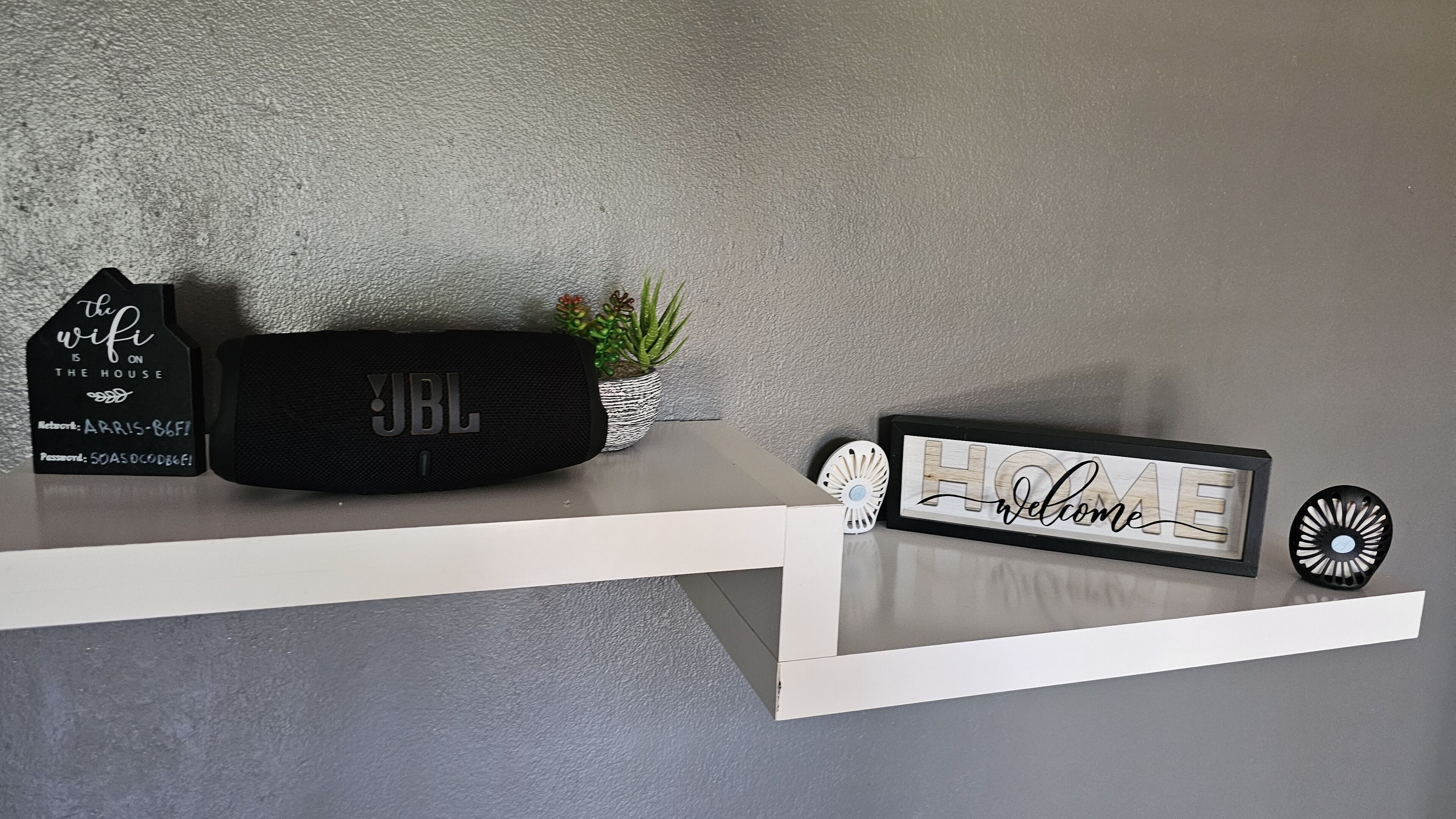 Een plankje met een zwarte JBL speaker, een bordje met de tekst 'wifi is gratis', een kleine potplant en een welkomstbord.