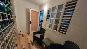 A room with a wooden floor, a black wicker sofa, a small table, and a chair. There is a window with blinds and a door with a metal gate. - Cozyville in the Manor (Ocho Rios)