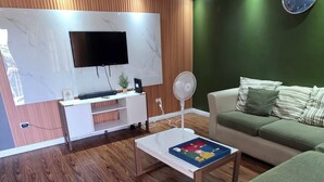 A modern living room with a wooden floor, a ceiling fan, a flat-screen TV mounted on a marble wall, a white entertainment unit, a green sofa, and a round clock. - Cozyville in the Manor (Ocho Rios)