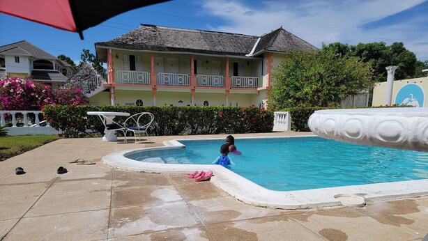 A swimming pool with a tiled deck, a house in the background, and a decorative fountain. - Cozyville in the Manor (Ocho Rios)