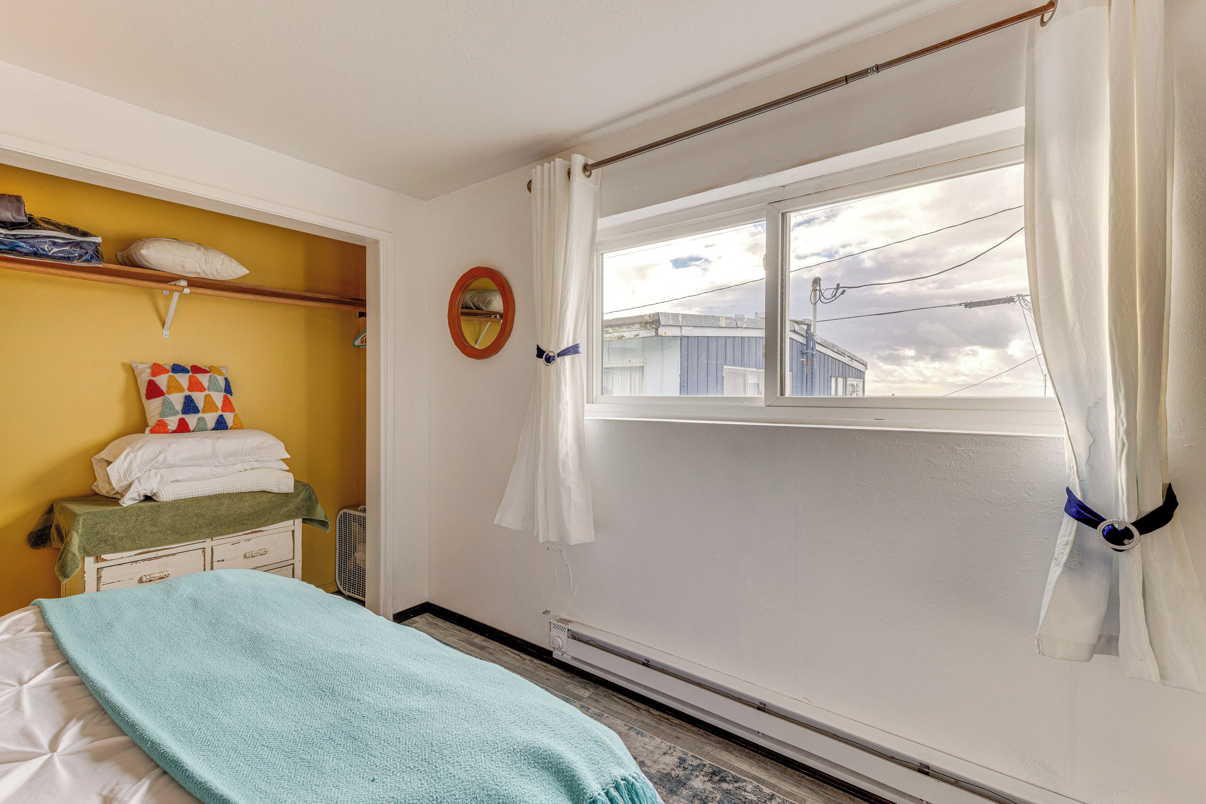 Une chambre à coucher avec un lit, une fenêtre avec des rideaux, un radiateur, un chauffage d’appoint, une étagère avec des serviettes pliées et une horloge murale.
