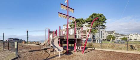 A playground with a slide and climbing structures, surrounded by a fence and a view of houses in the background.