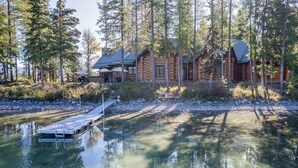A log cabin with a dock extending into a clear lake, surrounded by tall trees.