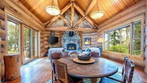 A log cabin interior with a stone fireplace, wooden dining table, and large windows.