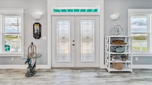 A foyer with a double glass door, a glass table with a sculpture, and a white shelving unit with decorative items.