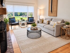 Ein Wohnzimmer mit Kamin, einer Couch, einem Couchtisch und Blick ins Freie.