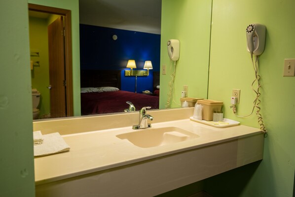 A hotel bathroom with a sink, mirror, hairdryer, and a bed visible in the reflection. - Budget Host Inn (Gilman)