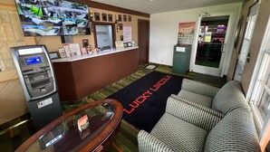 A lobby with an ATM, a reception desk, a large TV displaying security footage, a 'Lucky Lincoln' slot machine, and a 'Lucky Luke' themed carpet. - Budget Host Inn (Gilman)
