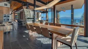 Une salle à manger spacieuse, avec une longue table et des chaises en bois, une cheminée en pierre et de grandes fenêtres offrant une vue sur la mer.