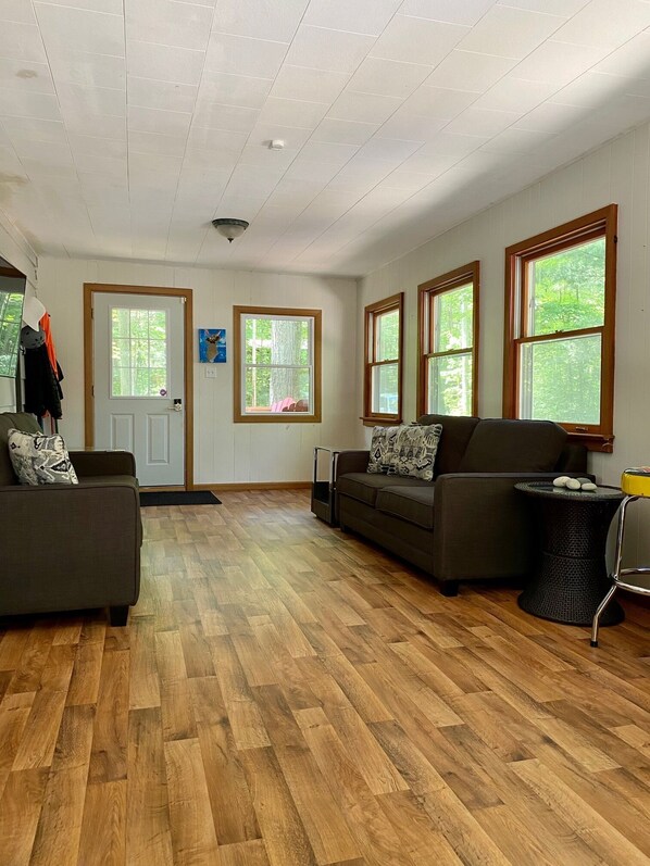 A spacious living room with hardwood flooring, two sofas, a coffee table, and large windows offering a view of greenery. - Updated Cottage in Southcott Pines, Grand Bend. Steps to Sun Beach (Grand Bend)