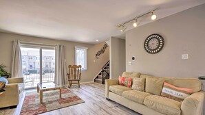 Ein Wohnzimmer mit einem beigen Sofa, einem Holztisch und einem gemusterten Teppich. Eine Treppe führt zu einer oberen Etage, und ein großes Fenster mit Vorhängen ist vorhanden.