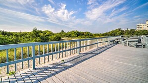 A wooden boardwalk with a blue railing, leading to a beach area with greenery and buildings in the background.