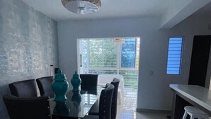 Uma sala de jantar moderna com mesa de vidro e cadeiras pretas, um candelabro e vista para o exterior através de uma janela ampla.