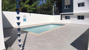Piscina com deck azulejado, cercada por um muro branco e palmeiras.