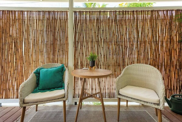 Two wicker chairs with cushions, a small round table, and a potted plant between them, all set on a wooden deck with a bamboo fence in the background.