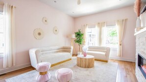 A modern living room with a white sofa, two pink ottomans, a round wooden coffee table, and a fireplace.