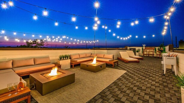 A rooftop terrace with outdoor seating, fire pits, and string lights against a twilight sky.