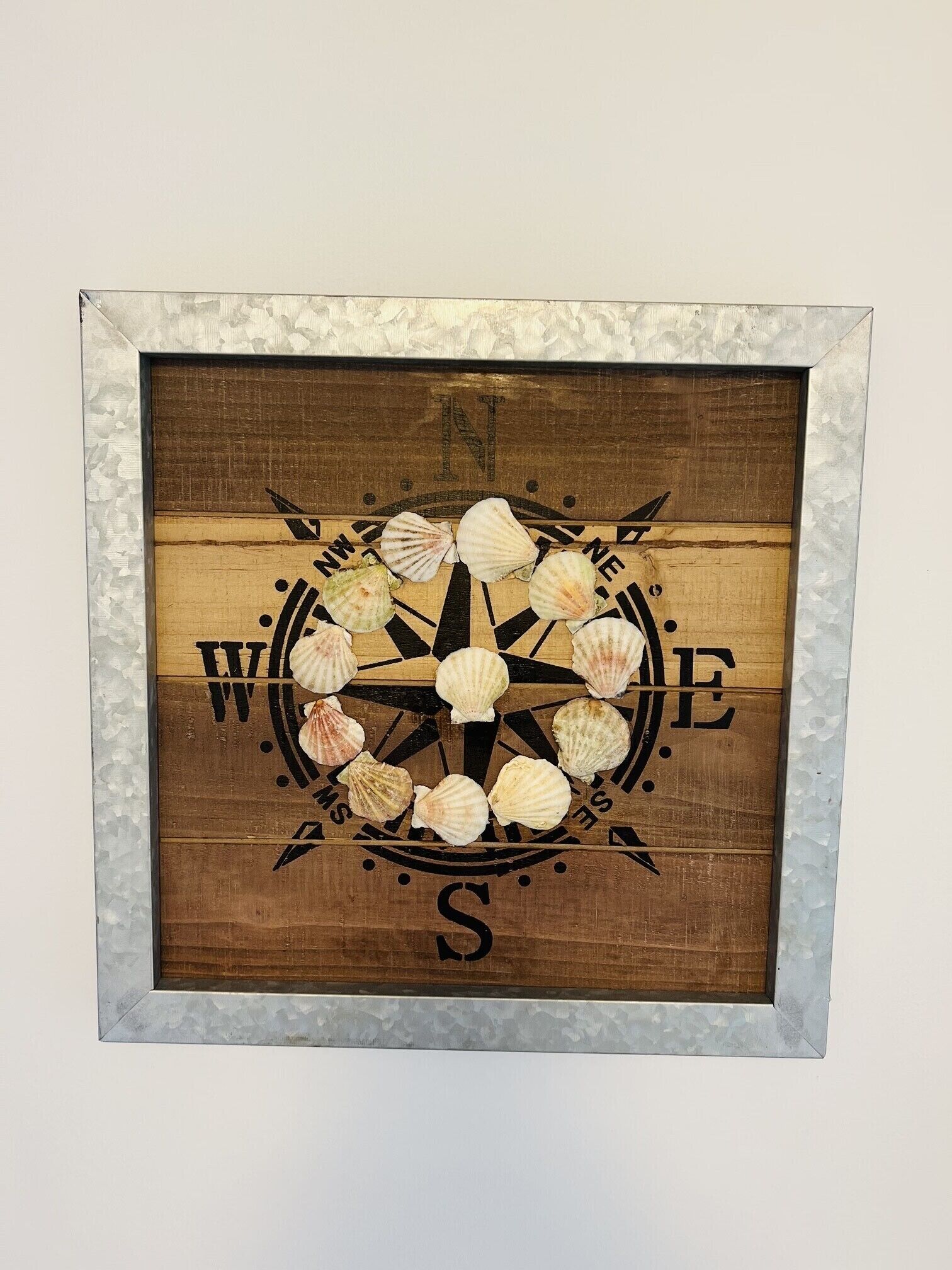 Ein gerahmter Kompass mit Muscheln und der Aufschrift 'WELCOME' und 'SOME' auf Holzuntergrund.