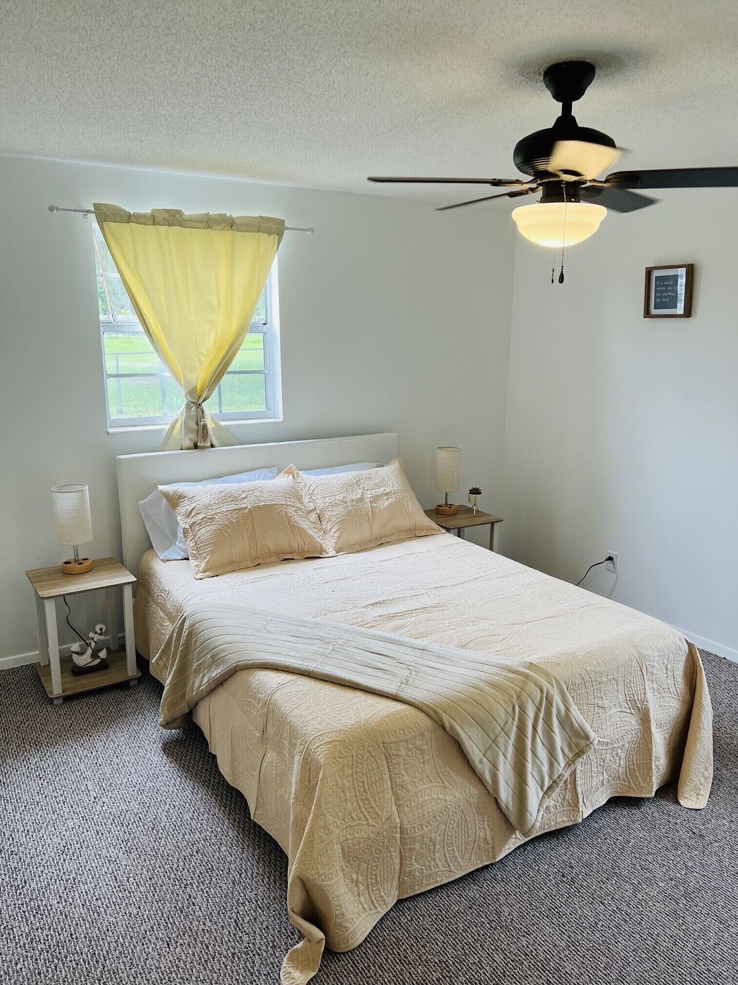 Ein Schlafzimmer mit Bett, Nachttisch, Lampe, Deckenventilator, Fenster mit Vorhängen und einem Wandbild.