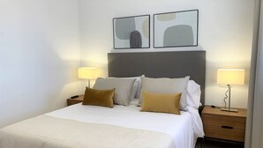 Un lit bien fait, avec une couette blanche et deux oreillers, flanqué de deux tables de chevet sur lesquelles reposent des lampes, dans une pièce dont le mur est d’un blanc uni et au-dessus du lit, on peut admirer une œuvre d’art abstraite.