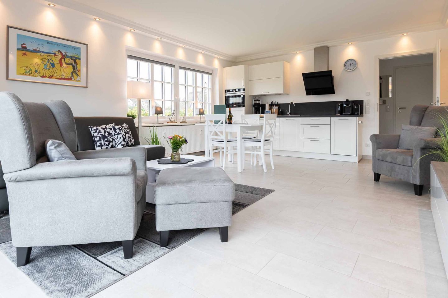 A modern living room with grey sofas, a dining area with white chairs, a kitchen with white cabinets, and a large window providing natural light.