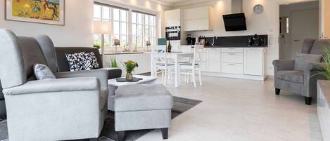 A modern living room with grey sofas, a dining area with white chairs, a kitchen with white cabinets, and a large window providing natural light.