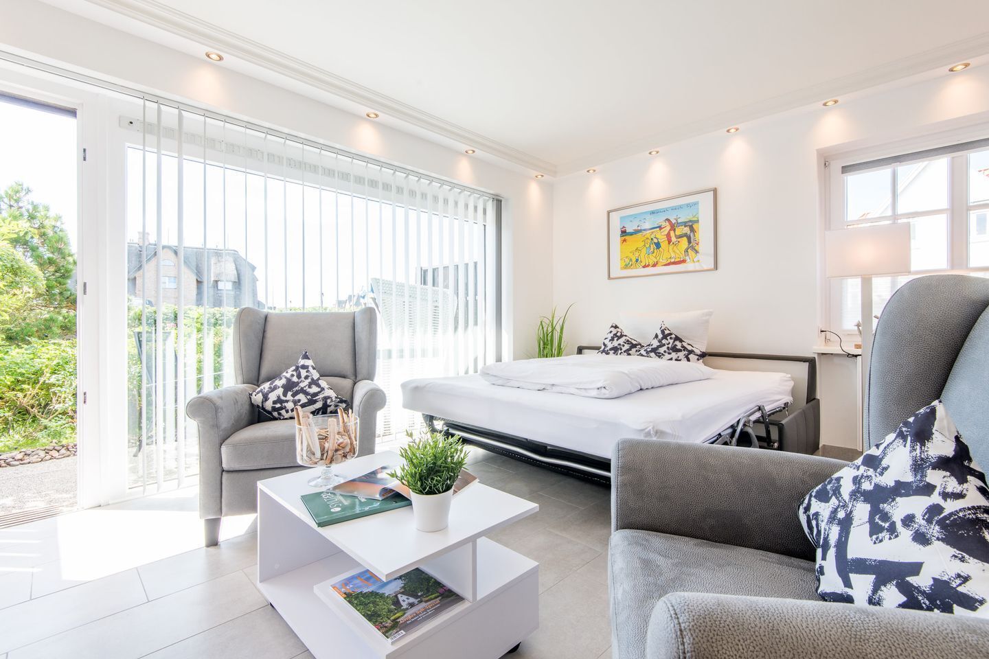 Un dormitorio moderno con cama, dos sillones, una mesita de centro y un amplio ventanal con persianas.