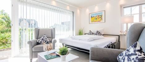 Un dormitorio moderno con cama, dos sillones, una mesita de centro y un amplio ventanal con persianas.