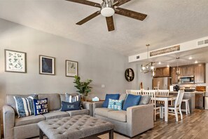 Un salon moderne avec un ensemble de canapés gris, un pouf capitonné et un ventilateur de plafond. On y trouve également un coin repas avec une table et des chaises blanches, ainsi qu’une cuisine dotée de meubles en bois et d’appareils électroménagers en acier inoxydable.