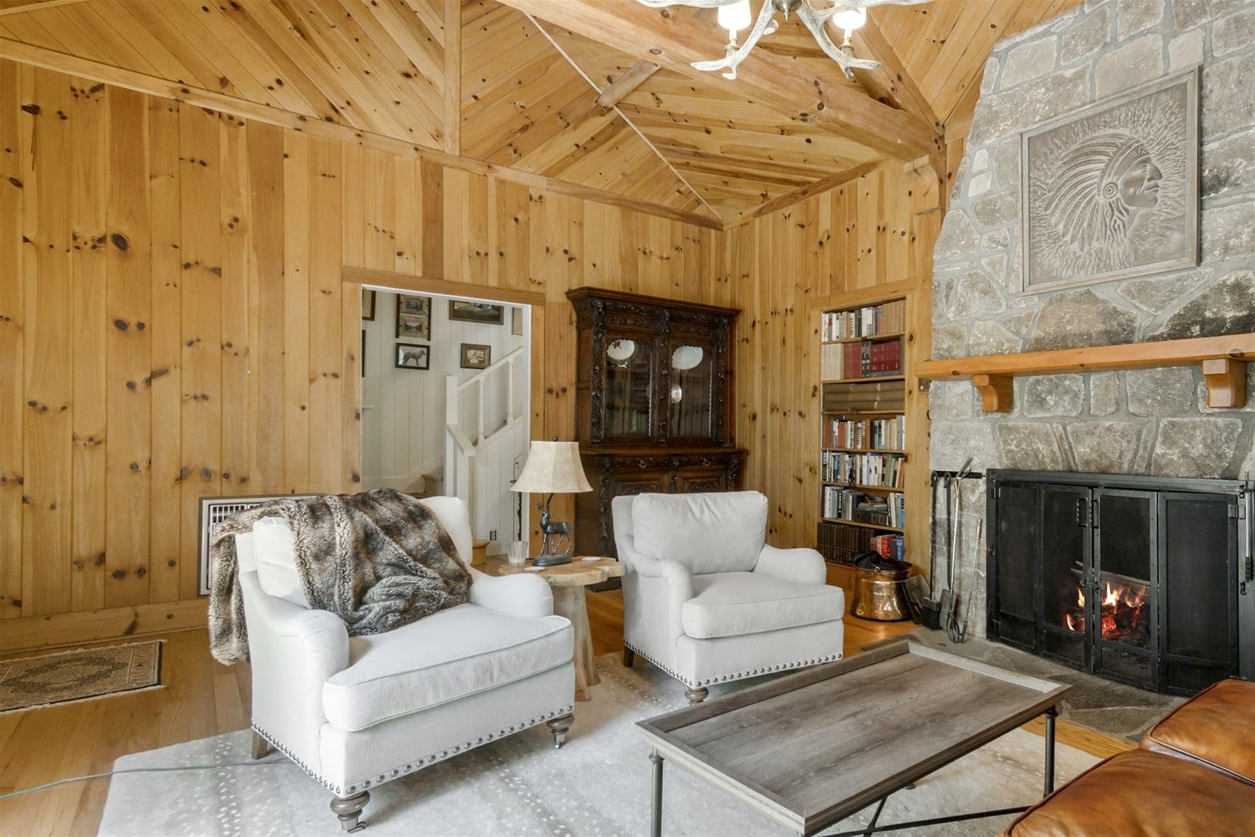 A cozy living room with wooden walls, two white armchairs, a fireplace, and a stone fireplace surround.