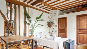 A rustic room with a wooden table, a bed with white linens, a blue ottoman, and a large plant. - Studio Listelli - Welkeys (Paris)