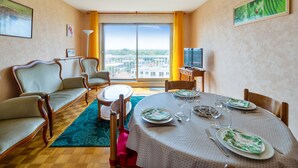 A hotel room with a dining area, a sofa, a television, and a large window with a city view. - Appartement Libération - Welkeys (Vincennes)