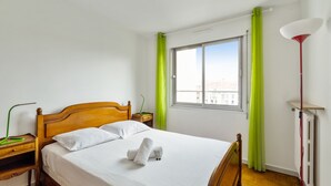 A neatly made bed with white linens and two pillows, a wooden nightstand with a green lamp, and a window with green curtains. - Appartement Libération - Welkeys (Vincennes)