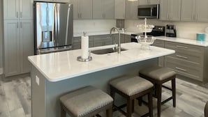Una cocina moderna con una isla central, electrodomésticos de acero inoxidable, iluminación colgante y diseño de armarios grises.