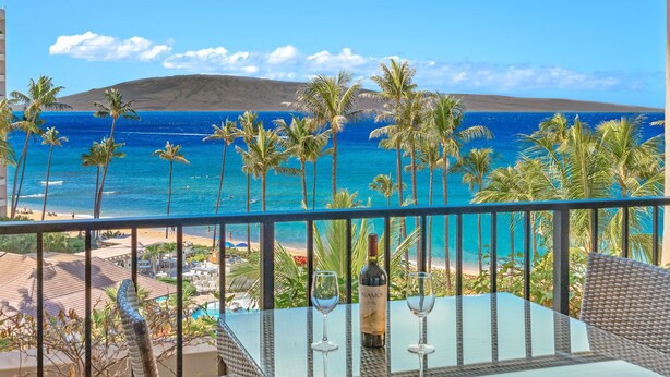 A balcony with a view of a beach, palm trees, and a bottle of wine.