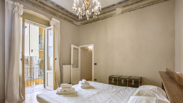 A bedroom with a bed, a bench, a chandelier, a suitcase, and a door leading to a balcony. - Myosotis - Sweet Dreams in Florence (Firenze)